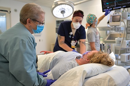 Two staff members in an ED room tend to a patient who is lying down, while in the background another staff member checks the readout on a monitor.
